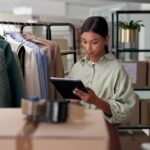 A woman in a clothing warehouse checks a tablet beside a rack of clothes.