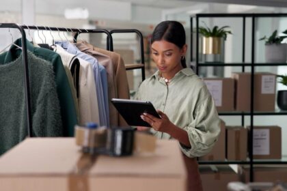 A woman in a clothing warehouse checks a tablet beside a rack of clothes.