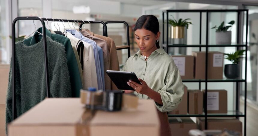 A woman in a clothing warehouse checks a tablet beside a rack of clothes.
