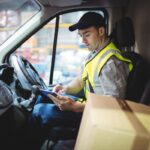A delivery driver sits in a van, reviewing information on a digital tablet.