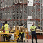 Warehouse workers in yellow safety vests push carts with boxes, surrounded by tall metal racks. A "DF ODD" sign hangs above them.