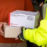 A postal worker in a bright yellow jacket hands two parcels to a person in an orange sweater. The scene conveys efficient mail delivery and service.