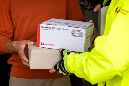 A postal worker in a bright yellow jacket hands two parcels to a person in an orange sweater. The scene conveys efficient mail delivery and service.