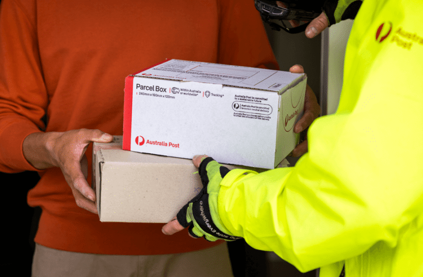 A postal worker in a bright yellow jacket hands two parcels to a person in an orange sweater. The scene conveys efficient mail delivery and service.