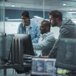 Three people work intently at a computer in a modern office. Monitors display code, suggesting a tech or programming environment. Collaborative atmosphere.