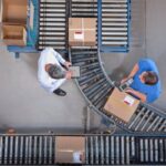 Two warehouse workers inspect packages on a conveyor system from an overhead perspective.