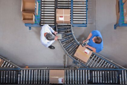 Two warehouse workers inspect packages on a conveyor system from an overhead perspective.