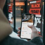 Person holding a smartphone displaying MediaMarkt store information in front of a Black Friday sign.