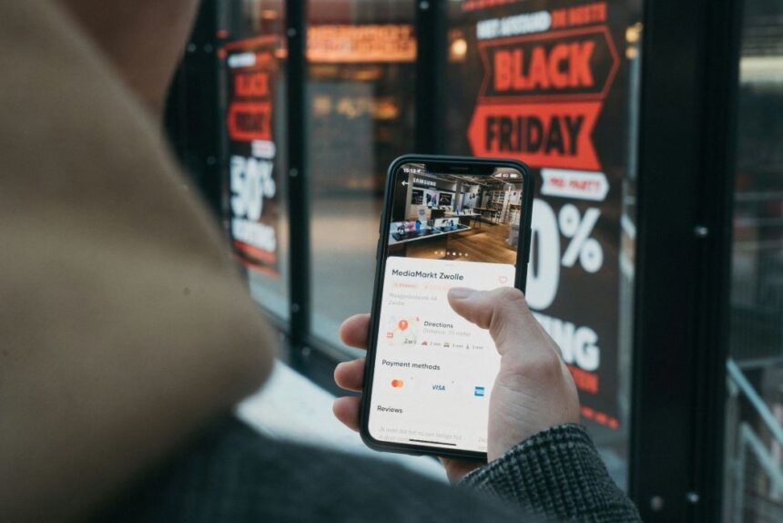 Person holding a smartphone displaying MediaMarkt store information in front of a Black Friday sign.