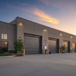 A modern industrial warehouse building with four roll-up garage doors at sunset.