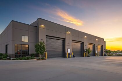 A modern industrial warehouse building with four roll-up garage doors at sunset.