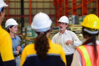 A manager leads a safety meeting with a team of industrial workers in a warehouse.