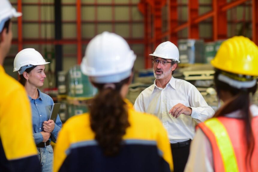 A manager leads a safety meeting with a team of industrial workers in a warehouse.