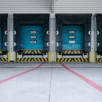 Four blue industrial loading dock doors with yellow safety stripes on a concrete lot.