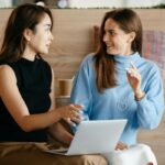 Two women sit together in a cafe, talking and looking at a laptop.