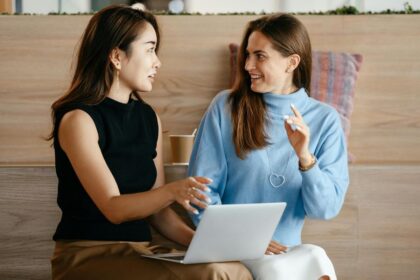 Two women sit together in a cafe, talking and looking at a laptop.