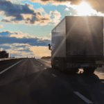 A semi-truck driving on a highway during a bright, cloudy sunset.