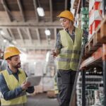 Warehouse workers in safety gear using a tablet to inspect inventory on shelves.