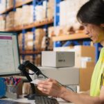 Warehouse worker scanning a package label in front of computer screens with tracking software.