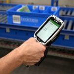 Handheld scanner showing warehouse stock picking software in front of blue conveyor belts.