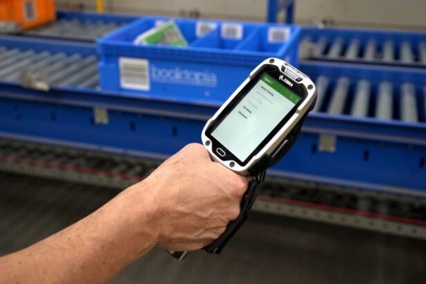 Handheld scanner showing warehouse stock picking software in front of blue conveyor belts.
