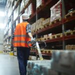 Warehouse worker in safety gear pulling a pallet jack through warehouse aisles.