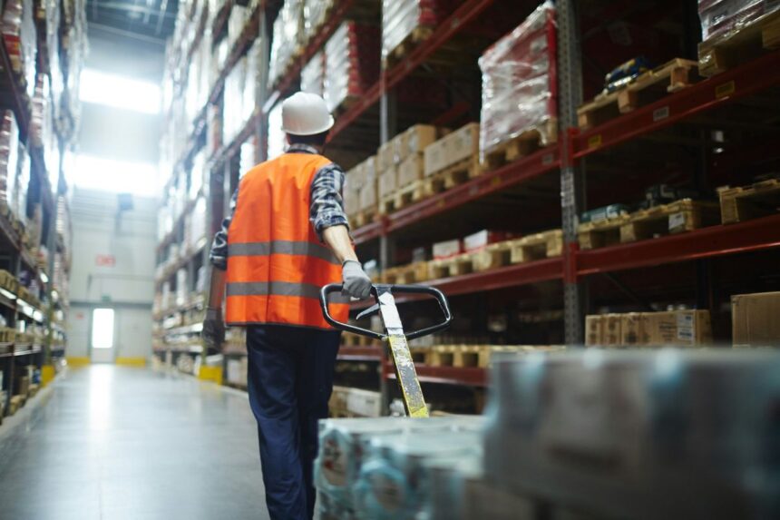Warehouse worker in safety gear pulling a pallet jack through warehouse aisles.