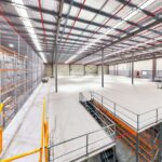 Large, modern industrial warehouse interior with mezzanine level, racking, and metal stairs.