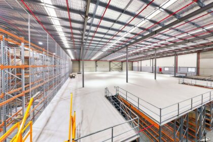 Large, modern industrial warehouse interior with mezzanine level, racking, and metal stairs.