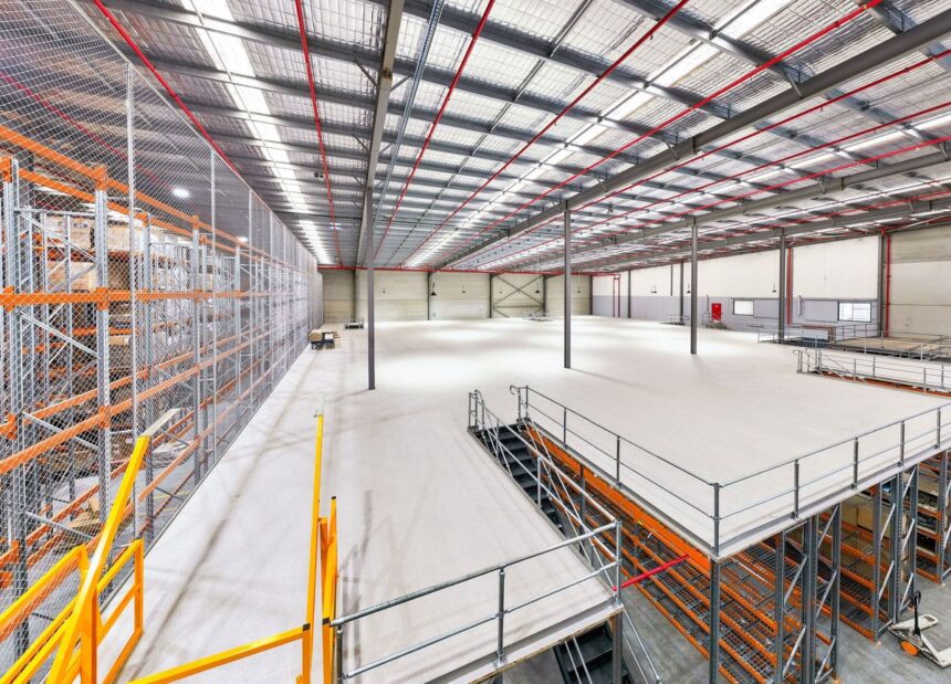 Large, modern industrial warehouse interior with mezzanine level, racking, and metal stairs.