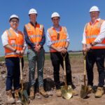 Four men in hard hats and high-visibility vests hold gold shovels at a construction site.