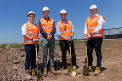 Four men in hard hats and high-visibility vests hold gold shovels at a construction site.
