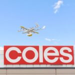 A delivery drone flying above a red Coles supermarket sign against a blue sky.