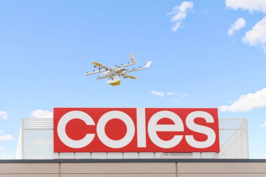 A delivery drone flying above a red Coles supermarket sign against a blue sky.
