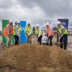 Six people in hard hats and vests performing a ceremonial groundbreaking at a construction site.
