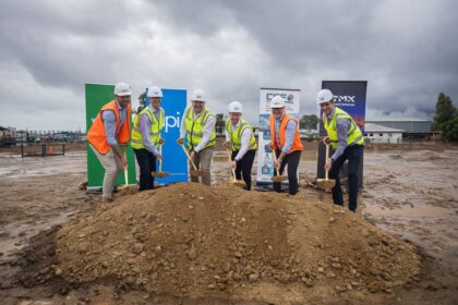 Six people in hard hats and vests performing a ceremonial groundbreaking at a construction site.