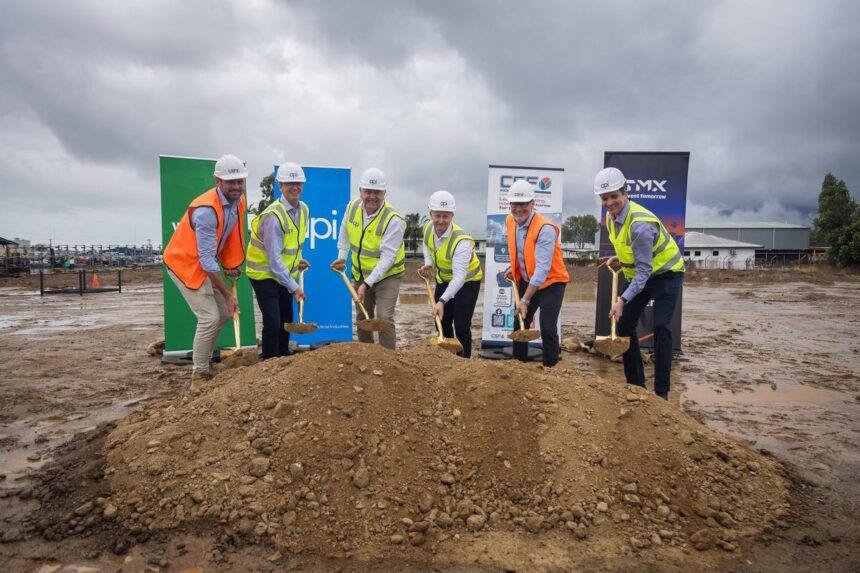 Six people in hard hats and vests performing a ceremonial groundbreaking at a construction site.