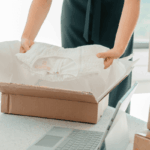 A person packing a white garment into a cardboard shipping box.