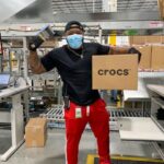 A warehouse worker in red pants holds a Crocs shipping box in a facility.