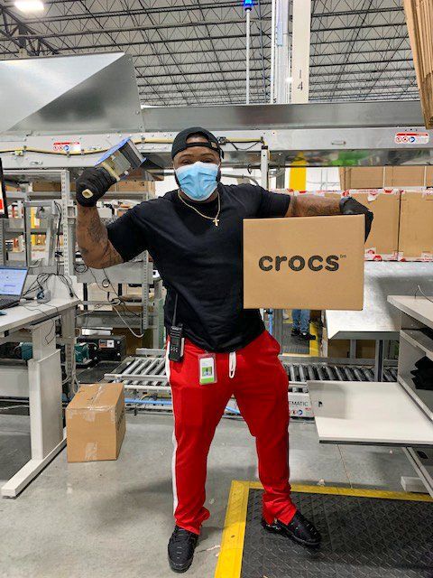 A warehouse worker in red pants holds a Crocs shipping box in a facility.