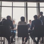 A group of professionals in business attire meeting in a bright, modern office boardroom.