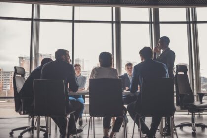 A group of professionals in business attire meeting in a bright, modern office boardroom.