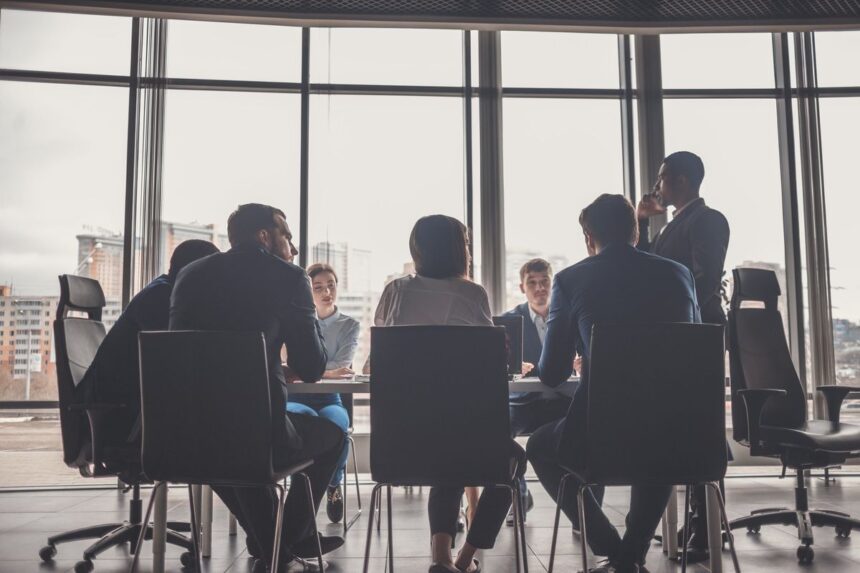 A group of professionals in business attire meeting in a bright, modern office boardroom.