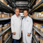 Two men in lab coats stand in a warehouse aisle between metal storage shelving.