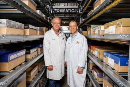 Two men in lab coats stand in a warehouse aisle between metal storage shelving.