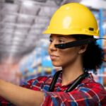 Warehouse worker wearing a yellow hard hat and smart glasses, holding a cardboard package.