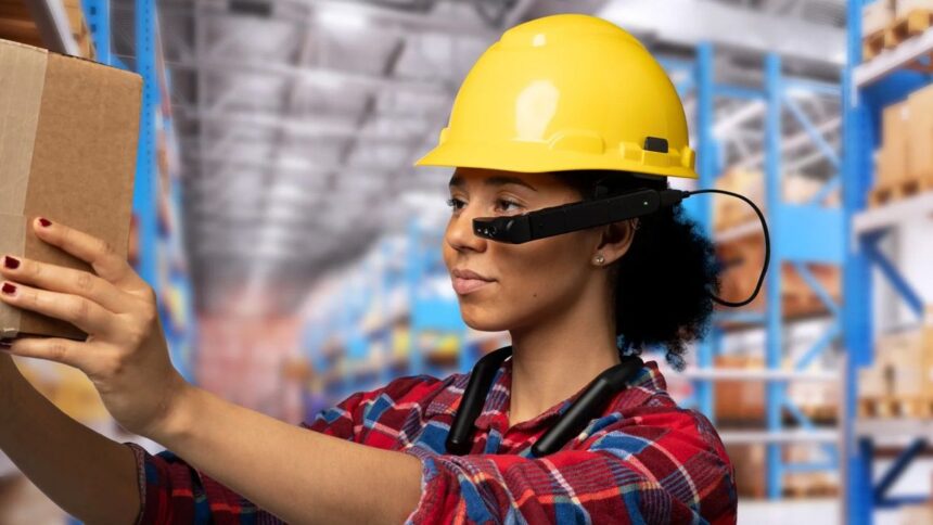 Warehouse worker wearing a yellow hard hat and smart glasses, holding a cardboard package.