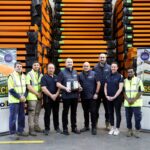A diverse team stands in a warehouse holding a Distributor of the Year trophy.