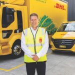 A DHL employee in a safety vest stands before electric DHL delivery vehicles.