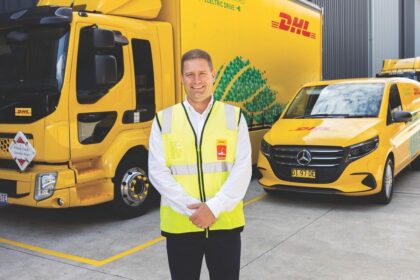 A DHL employee in a safety vest stands before electric DHL delivery vehicles.
